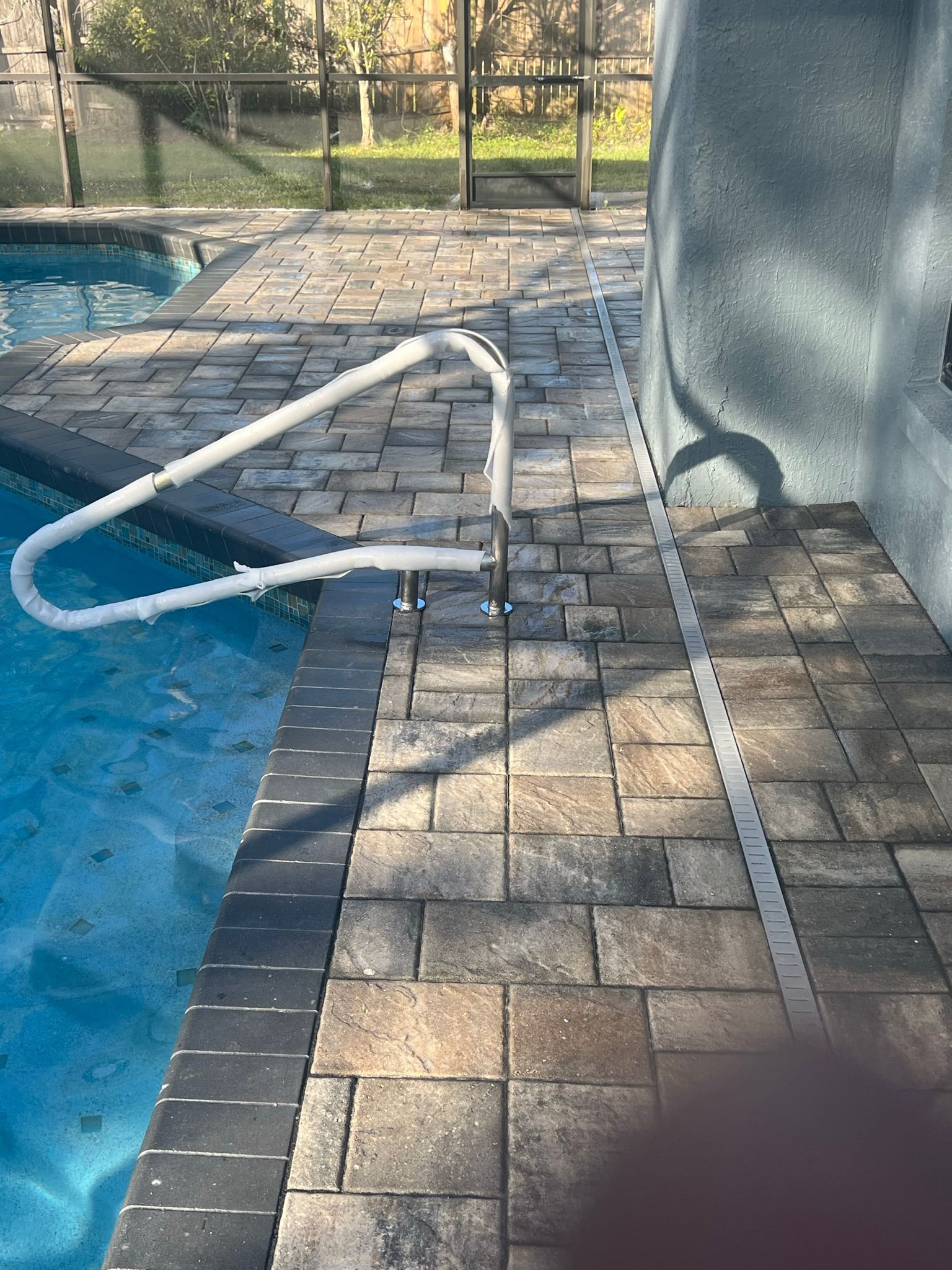 Poolside walkway with a white handrail, brick pavers, and a blue pool edge beside a gray wall.