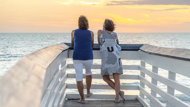 Two women are standing on a pier looking at the ocean at sunset.