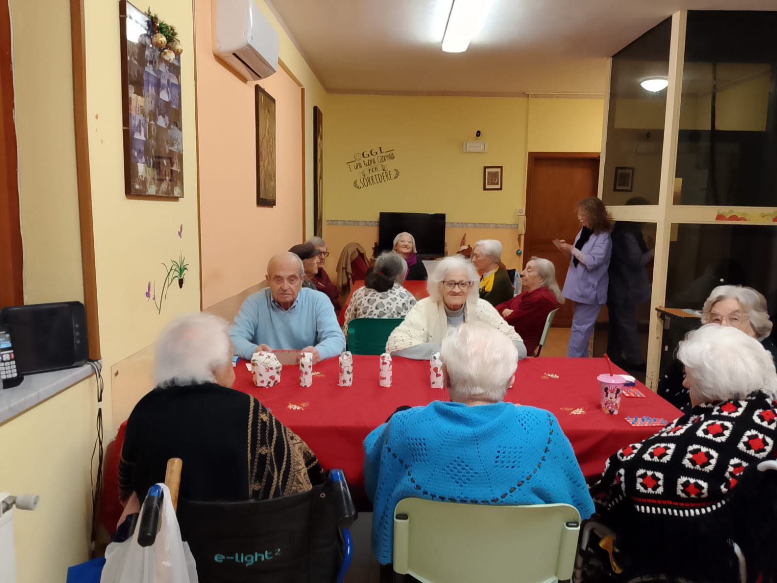Residenti che chiacchierano in un'accogliente sala comune attorno a una tovaglia rossa, con tazze e un televisore sullo sfondo.
