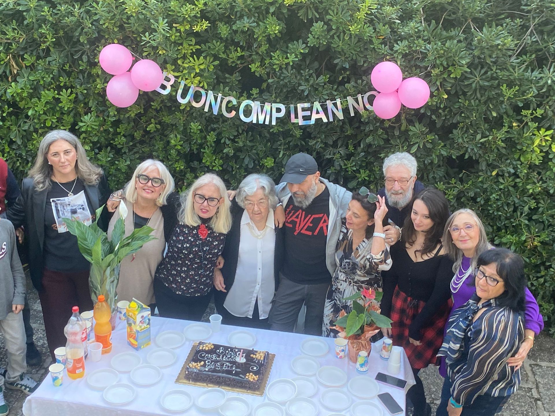 Le persone si sono riunite all'aperto intorno a una torta e a dei fiori per festeggiare, con palloncini rosa e uno striscione alle loro spalle.