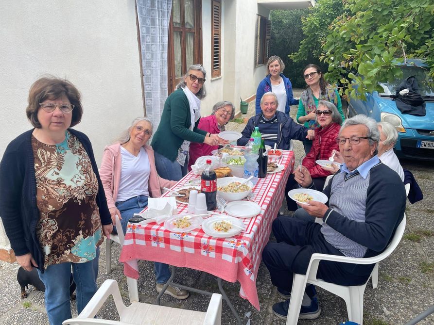 Un'assistente domiciliare serve il tè a un'anziana signora seduta a un tavolo con pasticcini e frutta in una stanza luminosa.