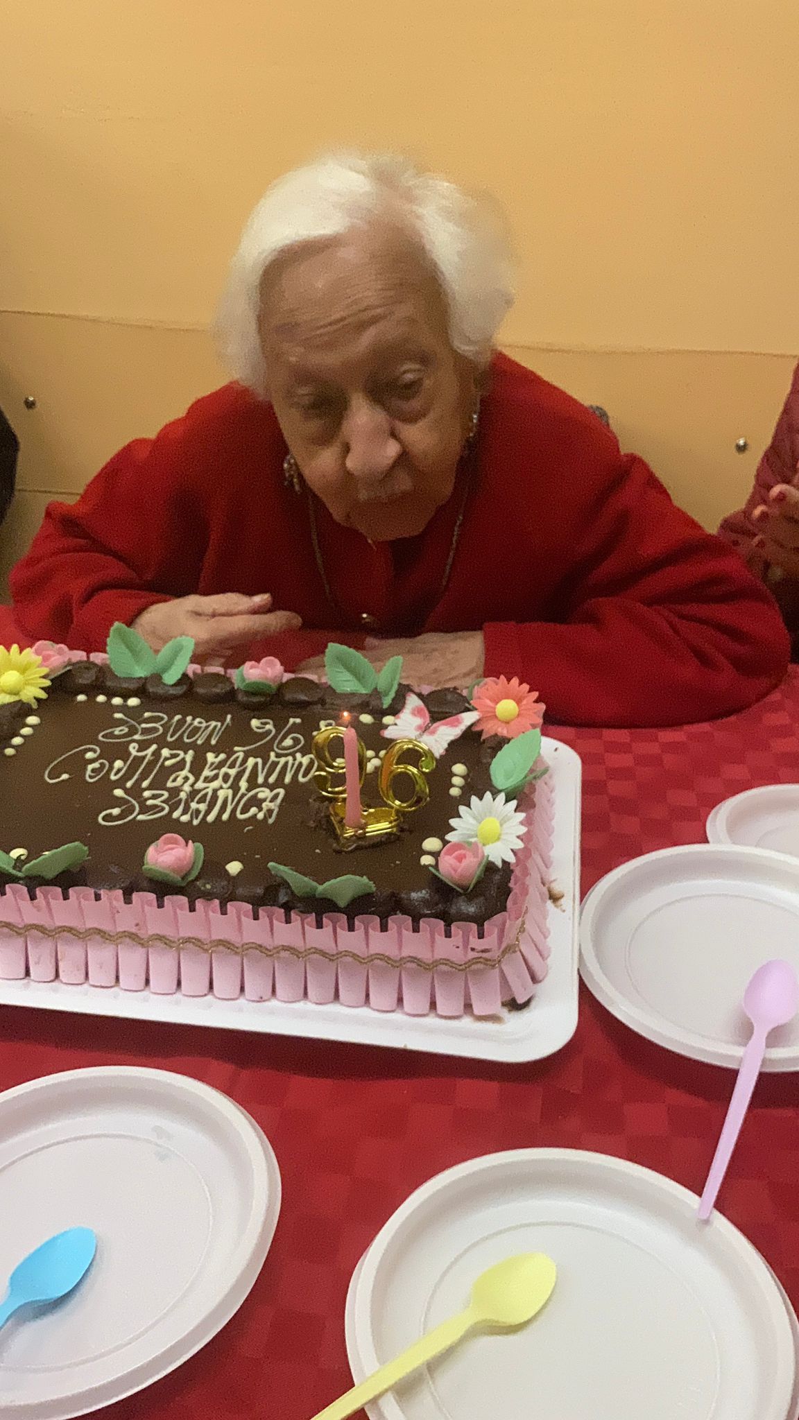 Una persona anziana con un maglione rosso spegne le candeline su una torta di compleanno al cioccolato seduta a un tavolo.