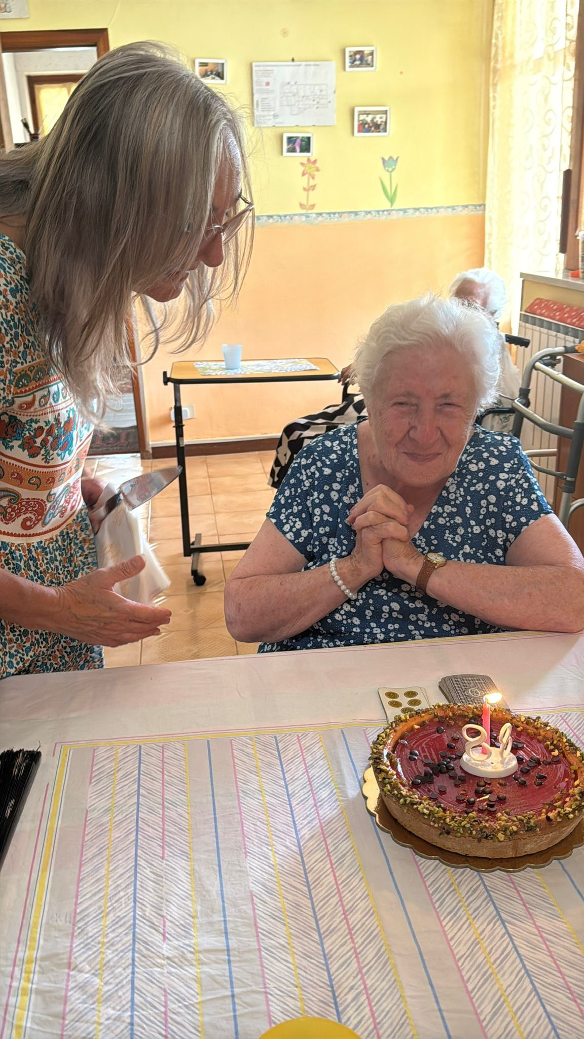 Una donna si china su una persona anziana accanto a una torta di compleanno illuminata, seduta a un tavolo in una stanza luminosa.