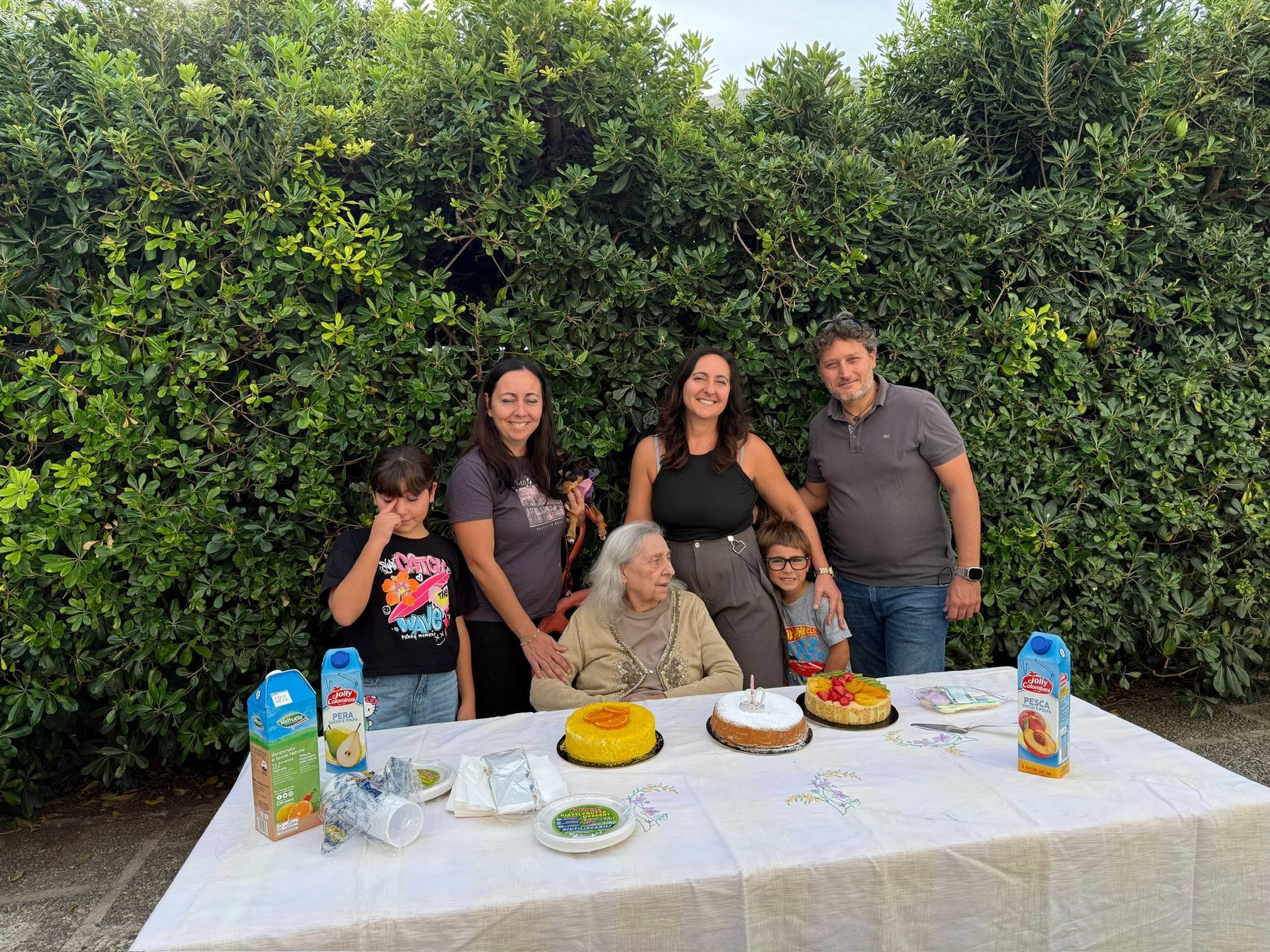 Una famiglia posa dietro un tavolo con torta, snack e bevande in un giardino sul retro.