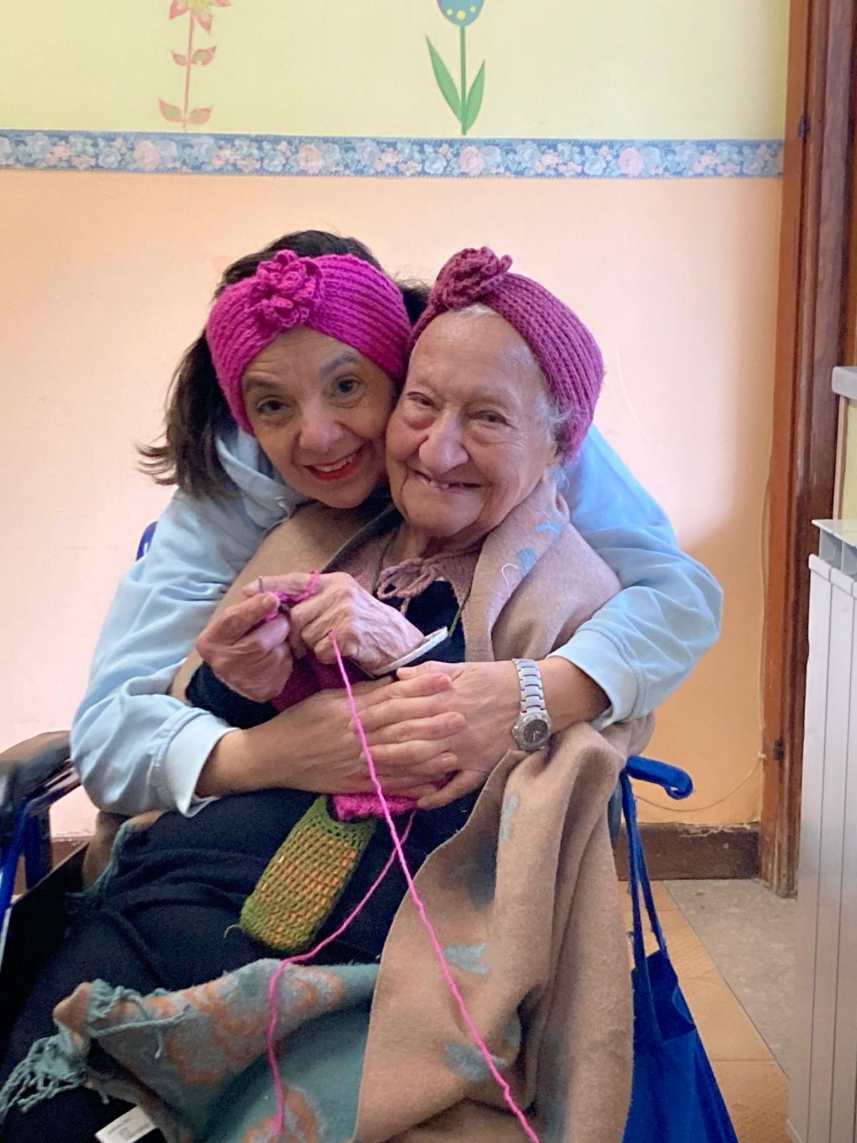 Due donne sorridenti e abbracciate in una stanza, entrambe con indosso cappelli di lana rosa, una delle quali tiene in mano un lavoretto artigianale realizzato con filato rosa.