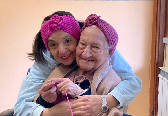 Due donne con fasce rosa per capelli si abbracciano e sorridono mentre lavorano a maglia insieme su un divano.