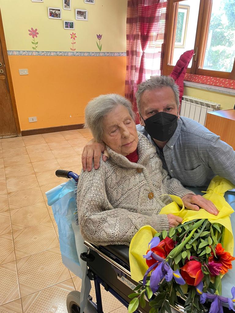 Due persone siedono su una sedia a rotelle all'interno di un edificio, una delle quali tiene in mano un colorato mazzo di fiori.