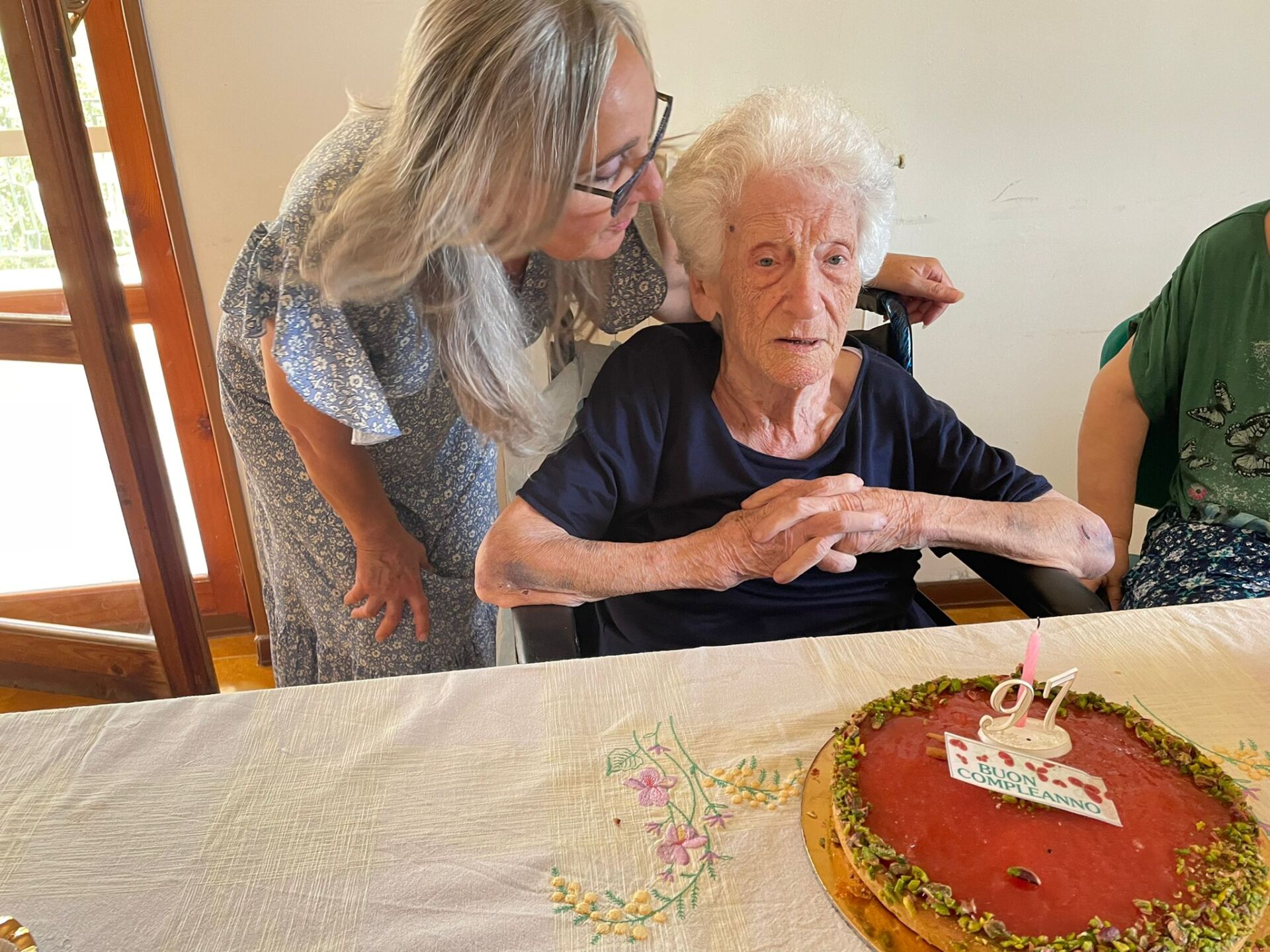 Una donna si china accanto a una persona anziana seduta a un tavolo con una torta di compleanno e una candelina accesa.