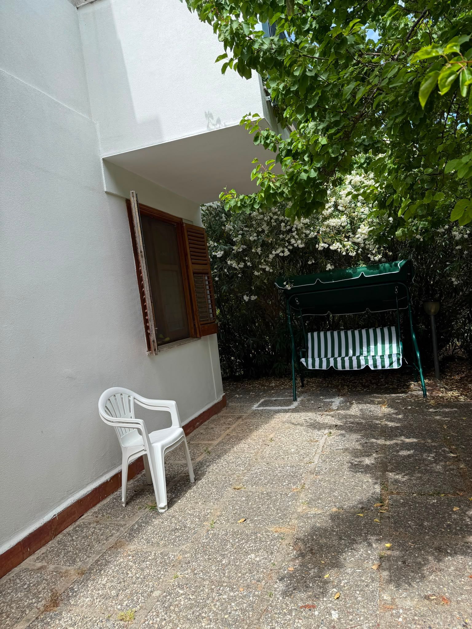 Cortile soleggiato accanto a un muro bianco, con una sedia bianca, pavimento in ghiaia e alberi ombrosi nelle vicinanze.