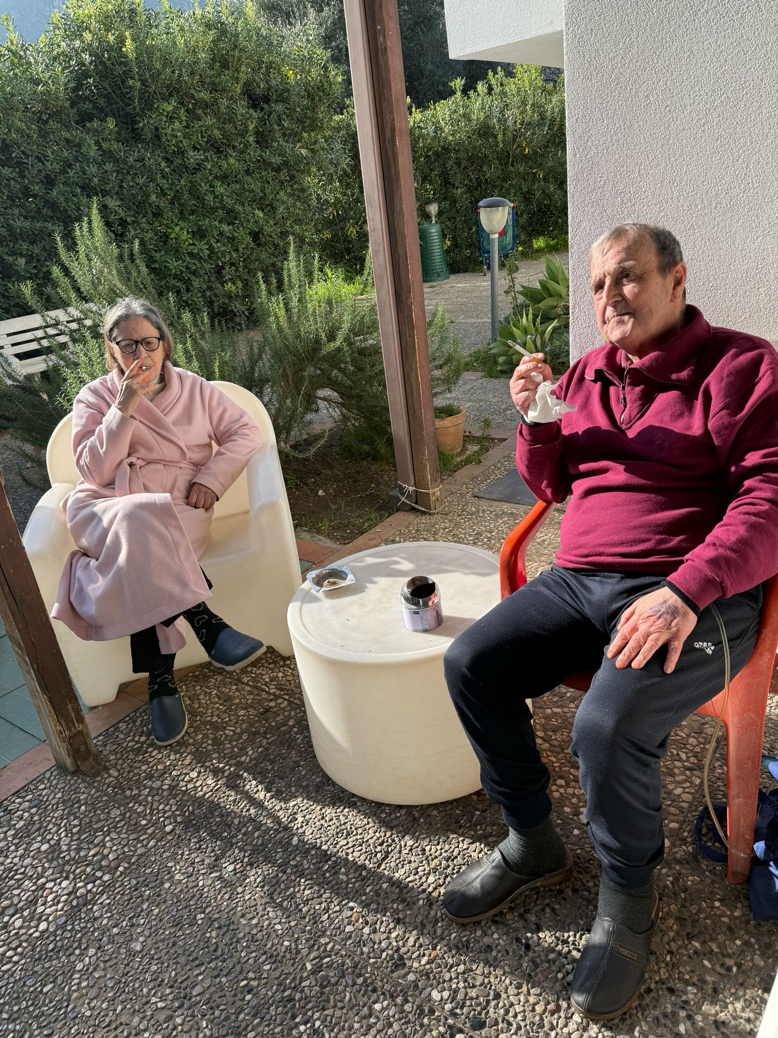 Due persone sedute all'aperto su delle sdraio accanto a un tavolino bianco in un giardino soleggiato.