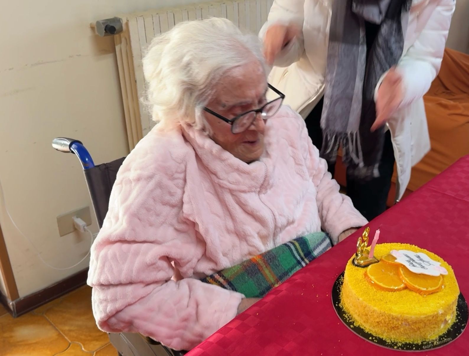 Una donna anziana con un maglione rosa guarda una piccola torta di compleanno gialla su una tovaglia rossa.