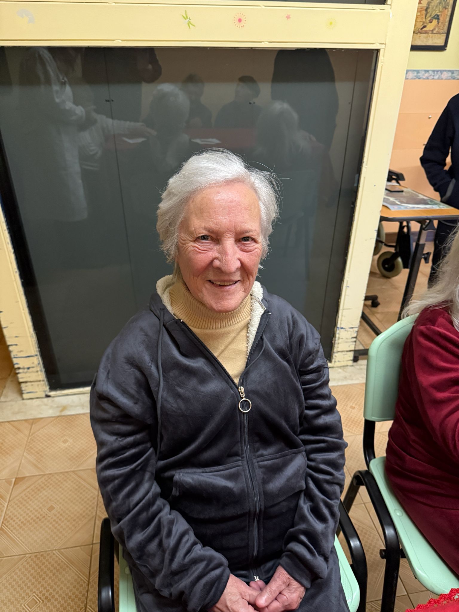 Donna sorridente con giacca nera seduta in un ambiente interno, appoggiata a una parete beige.