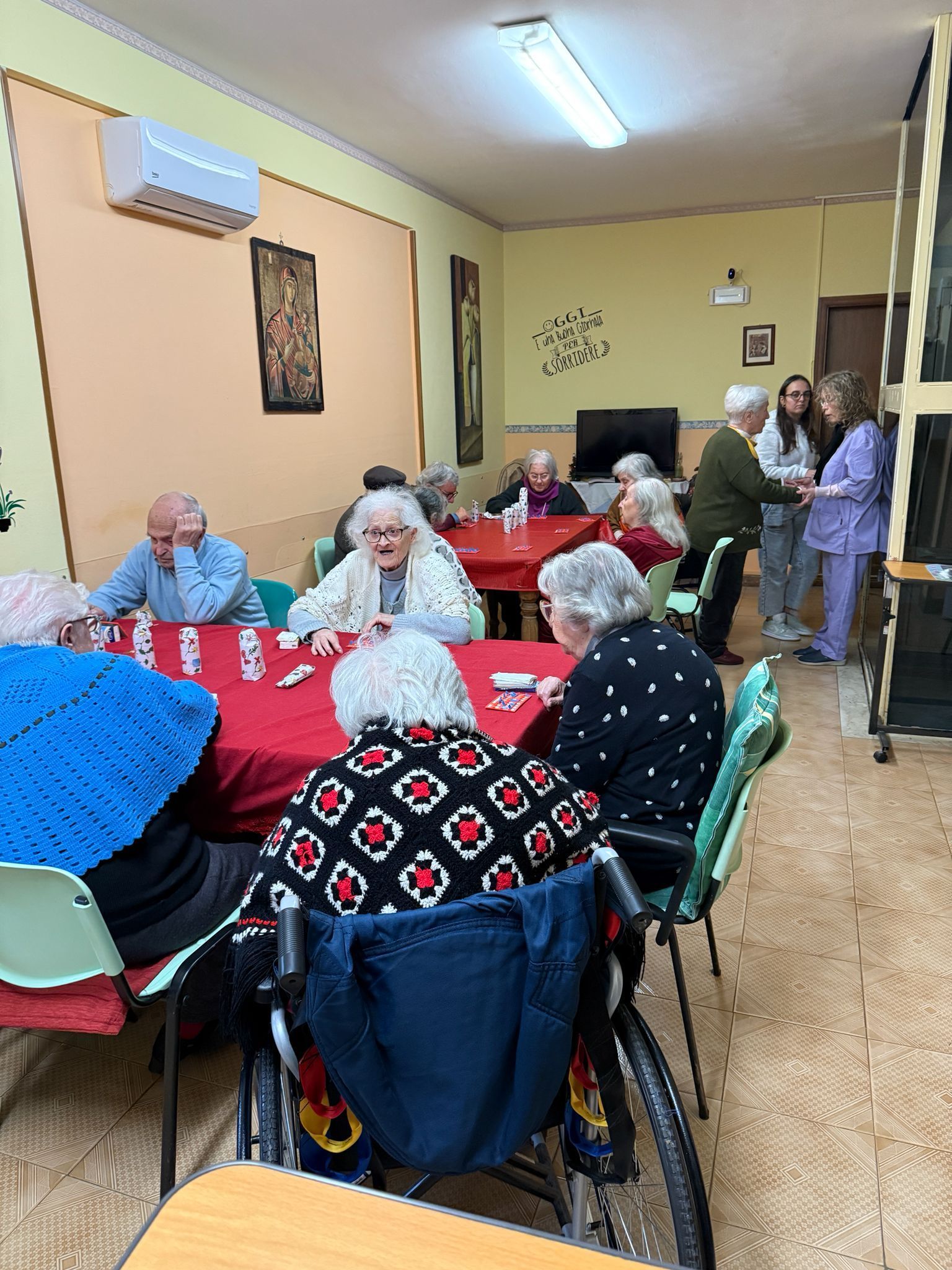 In una sala comune, le persone erano sedute attorno a tavoli ricoperti di tovaglie rosse, mentre altre erano in piedi vicino alla parete di fondo.