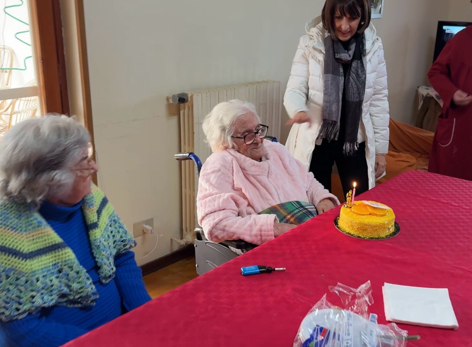 In una stanza, alcune persone sono riunite attorno a un tavolo con una torta di compleanno; una persona è in piedi accanto a due adulti seduti.