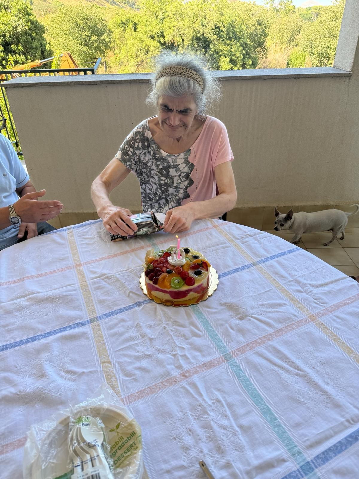 Una donna anziana taglia una torta a un tavolo all'aperto in un patio, con un cane sullo sfondo.