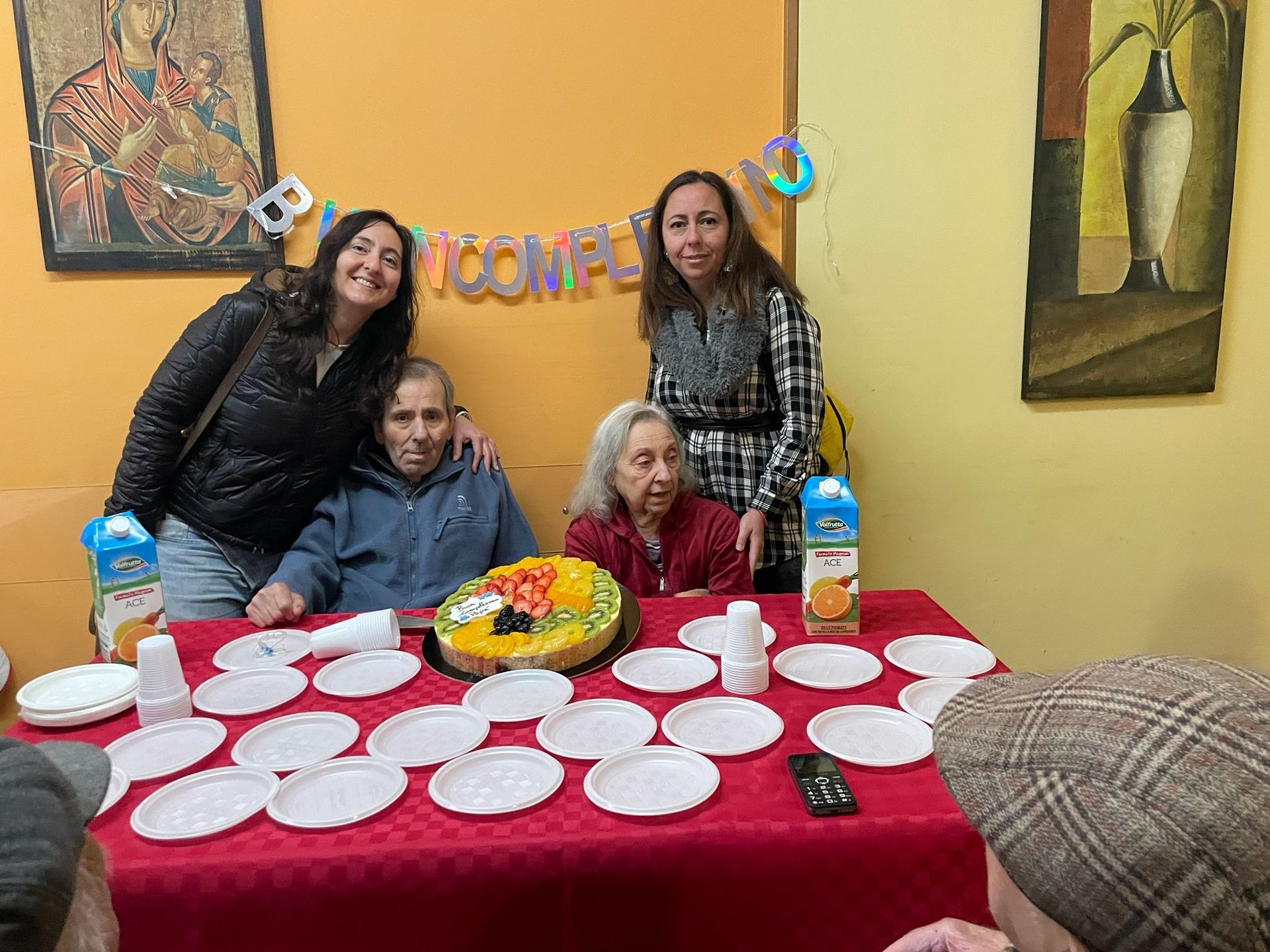 Un gruppo di quattro persone si è riunito attorno a un tavolo ricoperto da una tovaglia rossa, con piatti, torta e bevande, per una piccola festa.