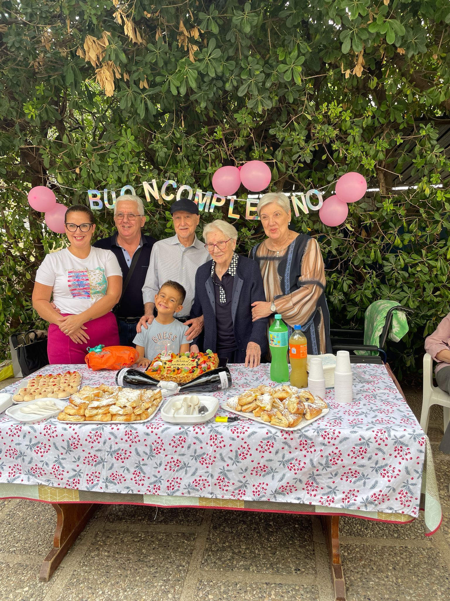 Gruppo di persone in posa dietro un tavolo di compleanno decorato con palloncini rosa e cibo all'aperto.