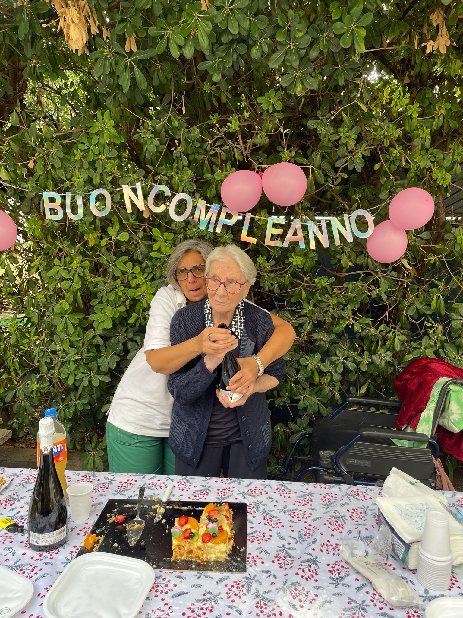 Due persone che si abbracciano a un tavolo da festa con torta, palloncini rosa e un cartello con la scritta 