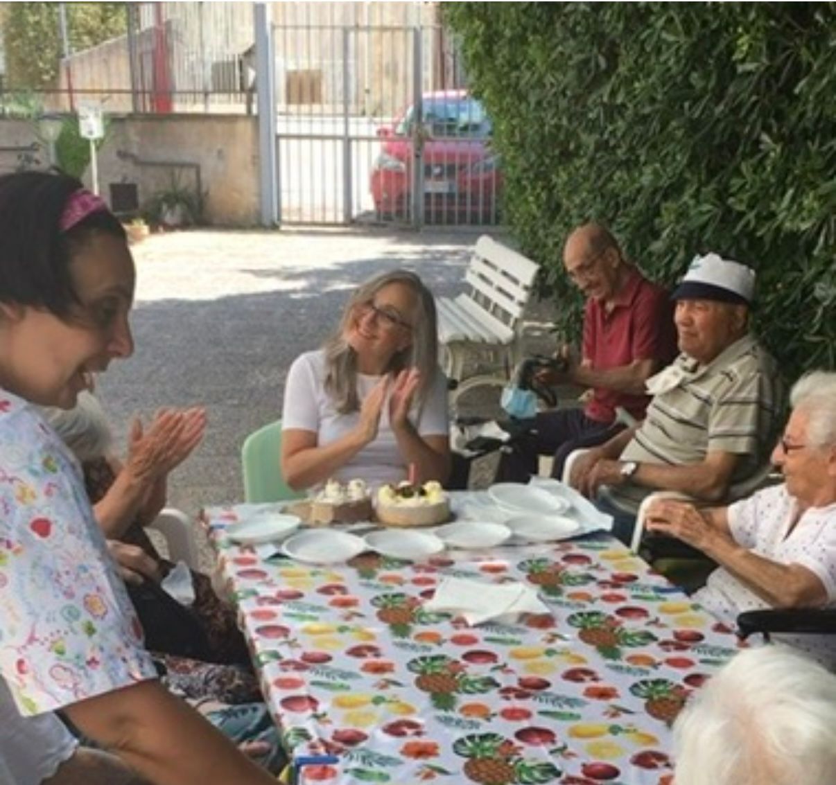 Persone sedute attorno a un tavolo durante un ritrovo all'aperto, con tazze, piatti e una tovaglia colorata.
