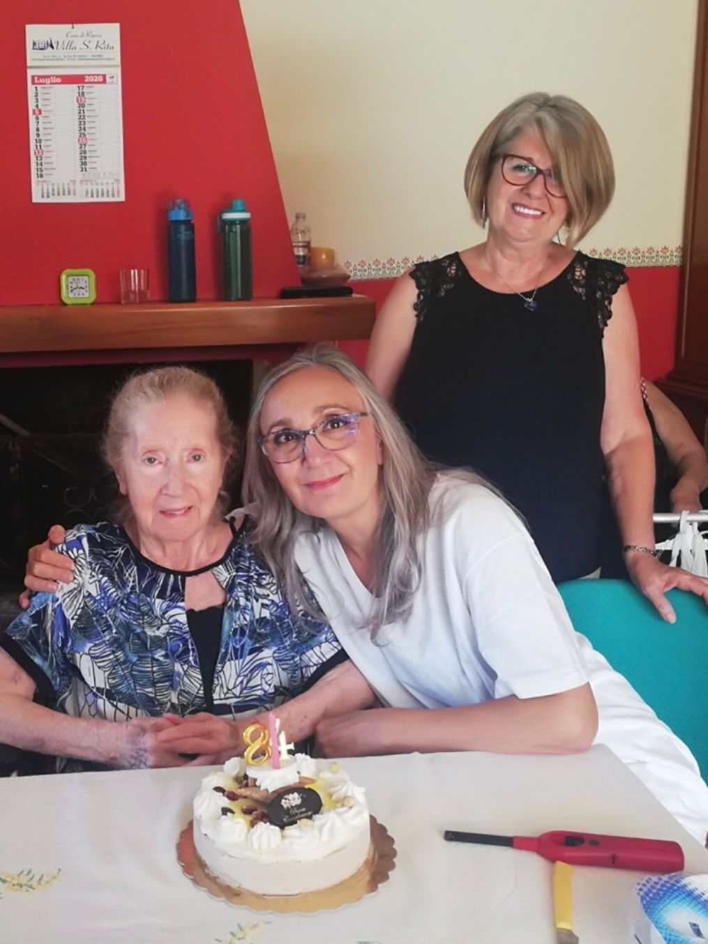 Tre donne sorridenti attorno a una torta di compleanno con una candelina accesa, sedute a un tavolo in una stanza rossa.