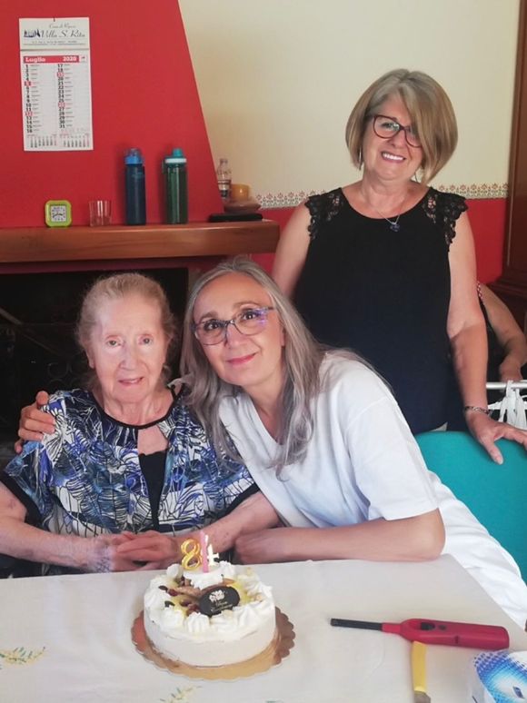 Un'assistente domiciliare sorride e conforta una donna seduta che tiene in mano una tazza e un libro in una luminosa sala comune di una casa di riposo.