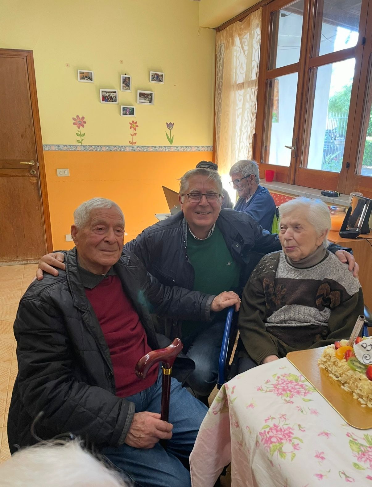 In una stanza luminosa, vicino a una finestra, tre persone siedono sorridenti a un tavolo.