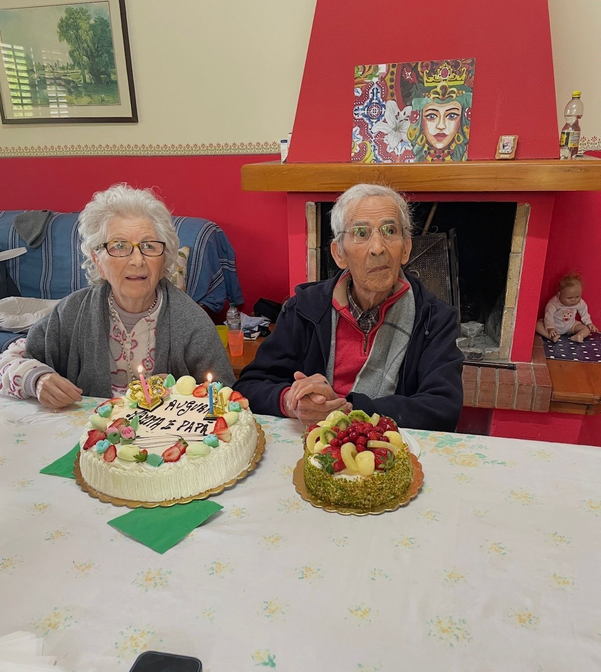 Due persone sedute a un tavolo con torte di compleanno, davanti a un camino rosso e a un ritratto incorniciato.