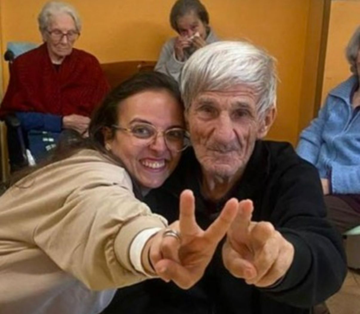 Una donna sorridente e un uomo anziano si sporgono in avanti e fanno il segno della pace in una stanza, con due persone sedute dietro di loro.
