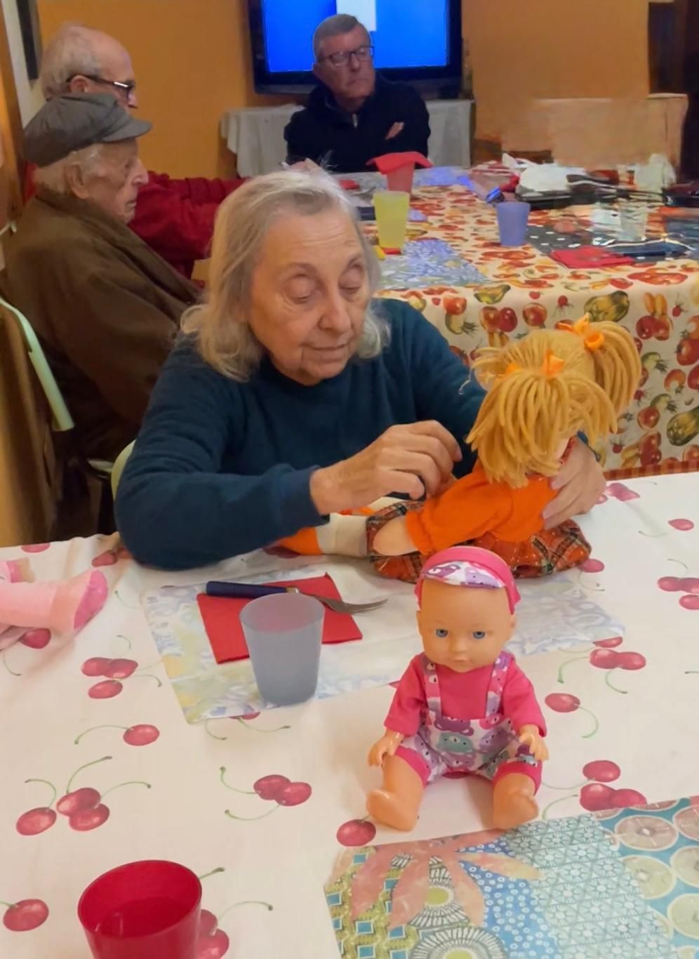 Un'anziana signora acconcia i capelli di una bambola a un tavolo durante un ricevimento festivo al chiuso.