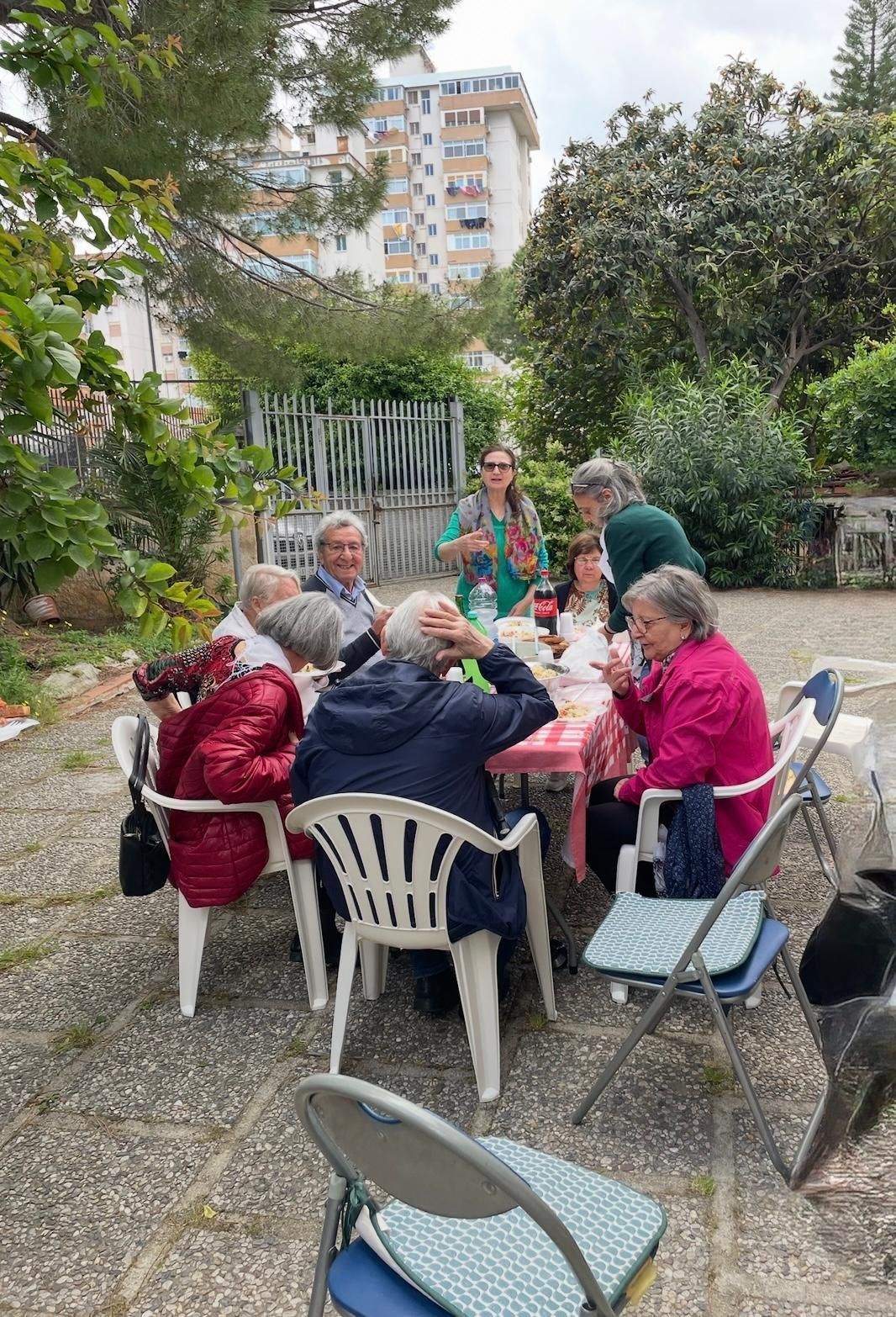 Persone sedute attorno a un tavolo all'aperto nel cortile di un giardino, chiacchierano e mangiano insieme.