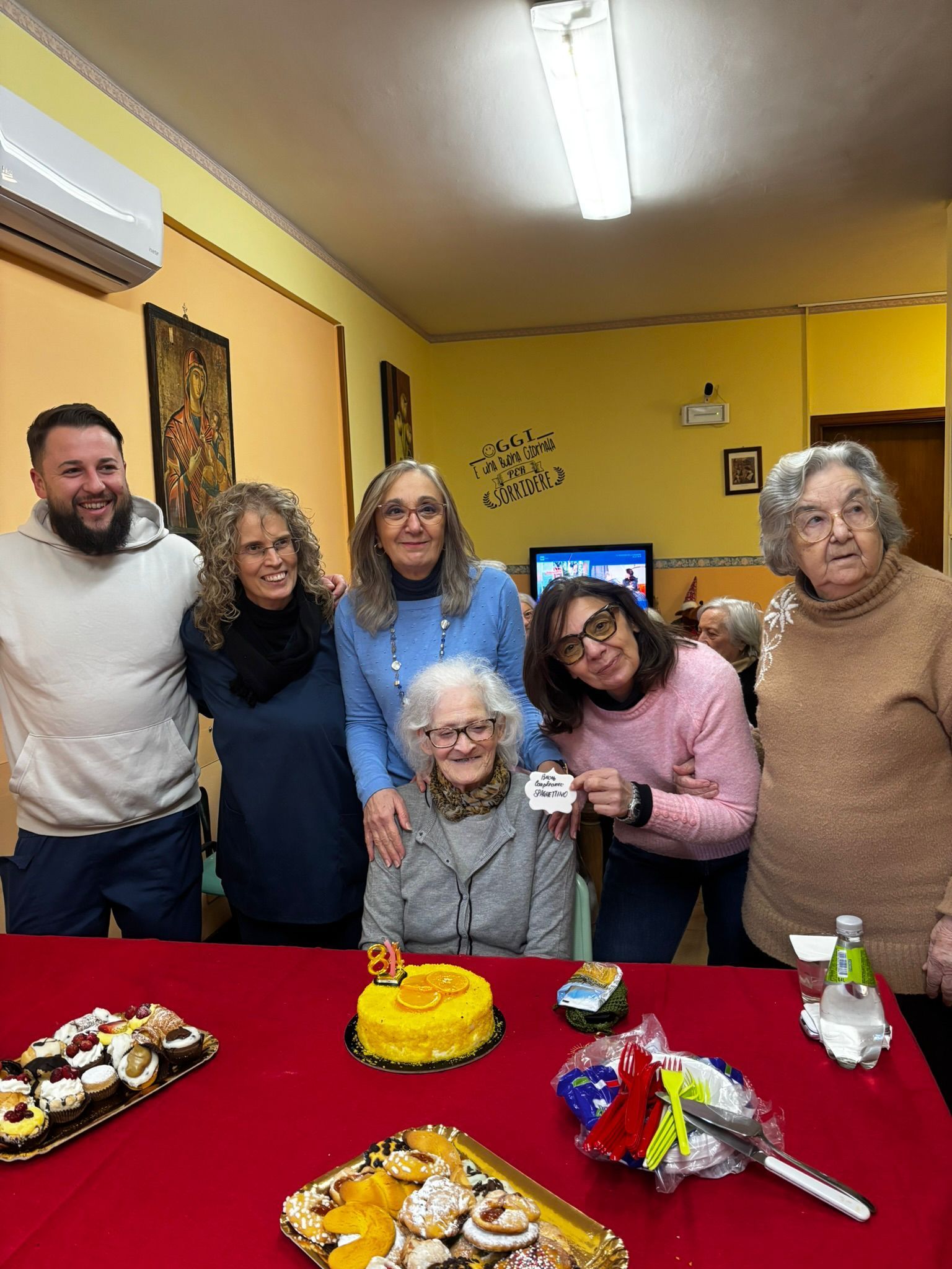 Un gruppo di persone sorridenti attorno a un tavolo con torte e dolcetti durante una festa di compleanno in una stanza gialla.