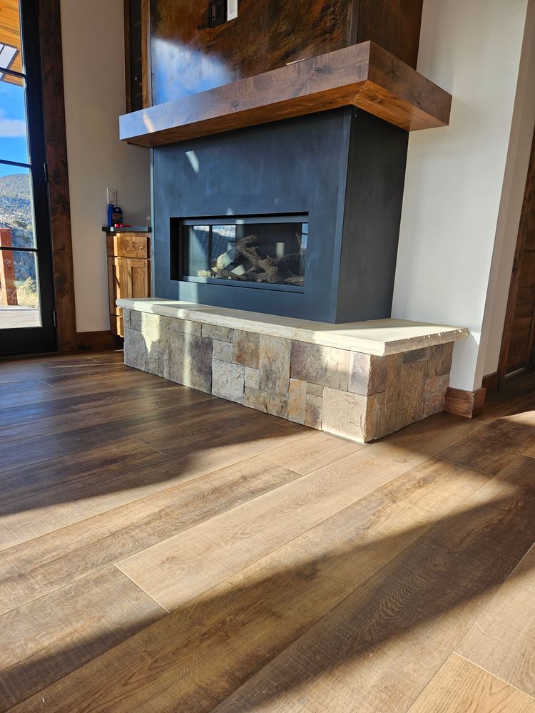 Fireplace with a stone base, dark gray surround, and wooden mantel in a room with wood floors.