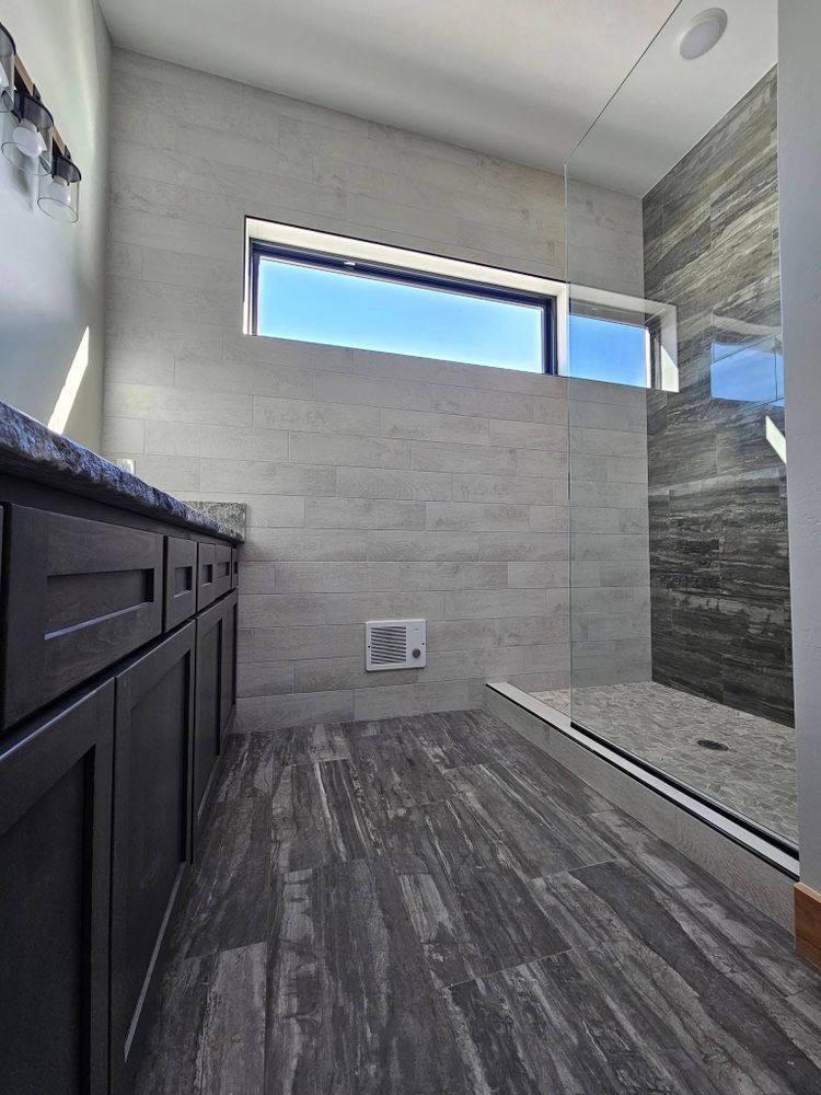 Modern bathroom with gray wood-look flooring and wall tiles, dark cabinets, and a glass shower.