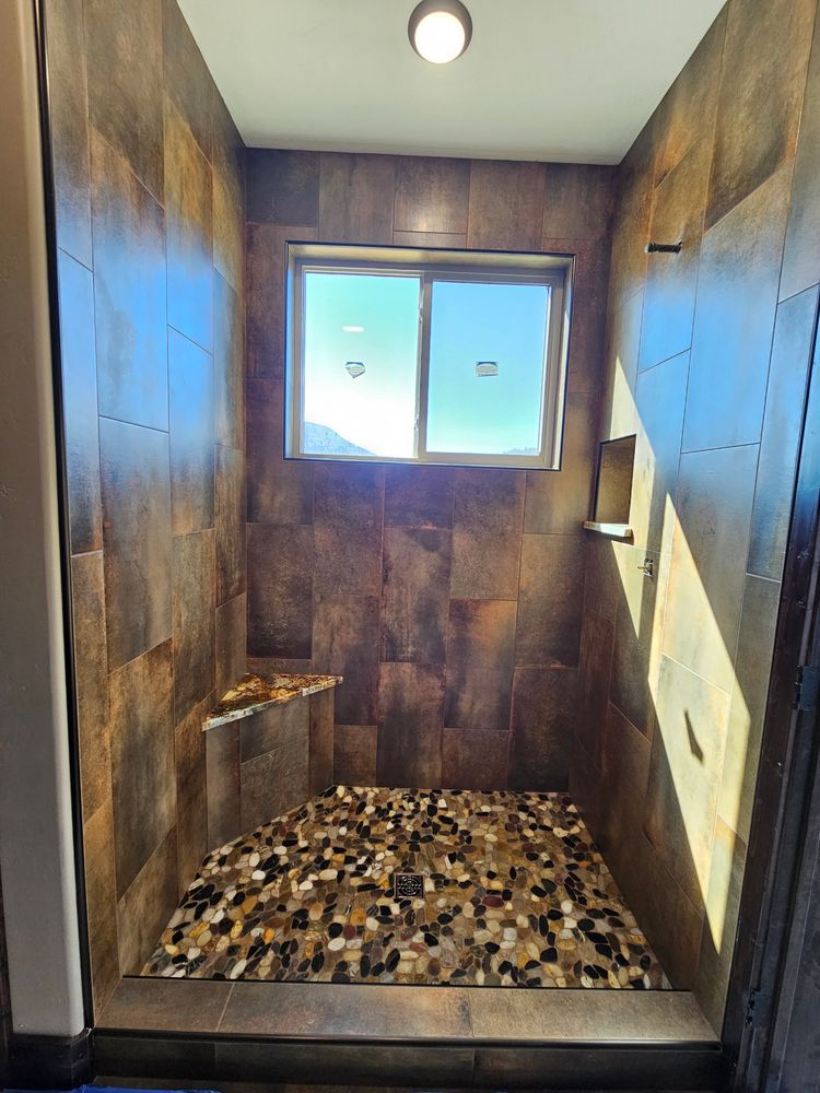 Shower with dark, textured tile walls, pebble floor, small window, and built-in soap ledges.
