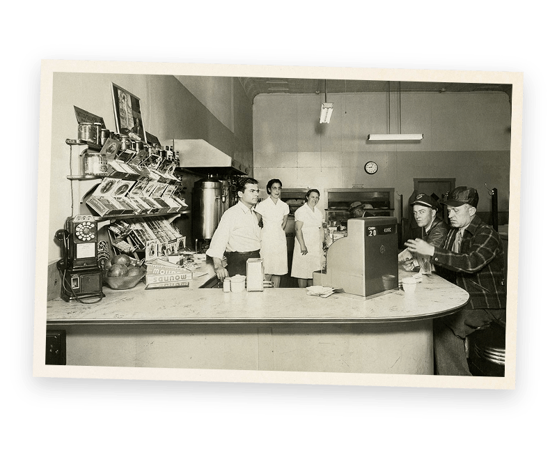 A black and white photo of a group of people standing around a counter in a restaurant.