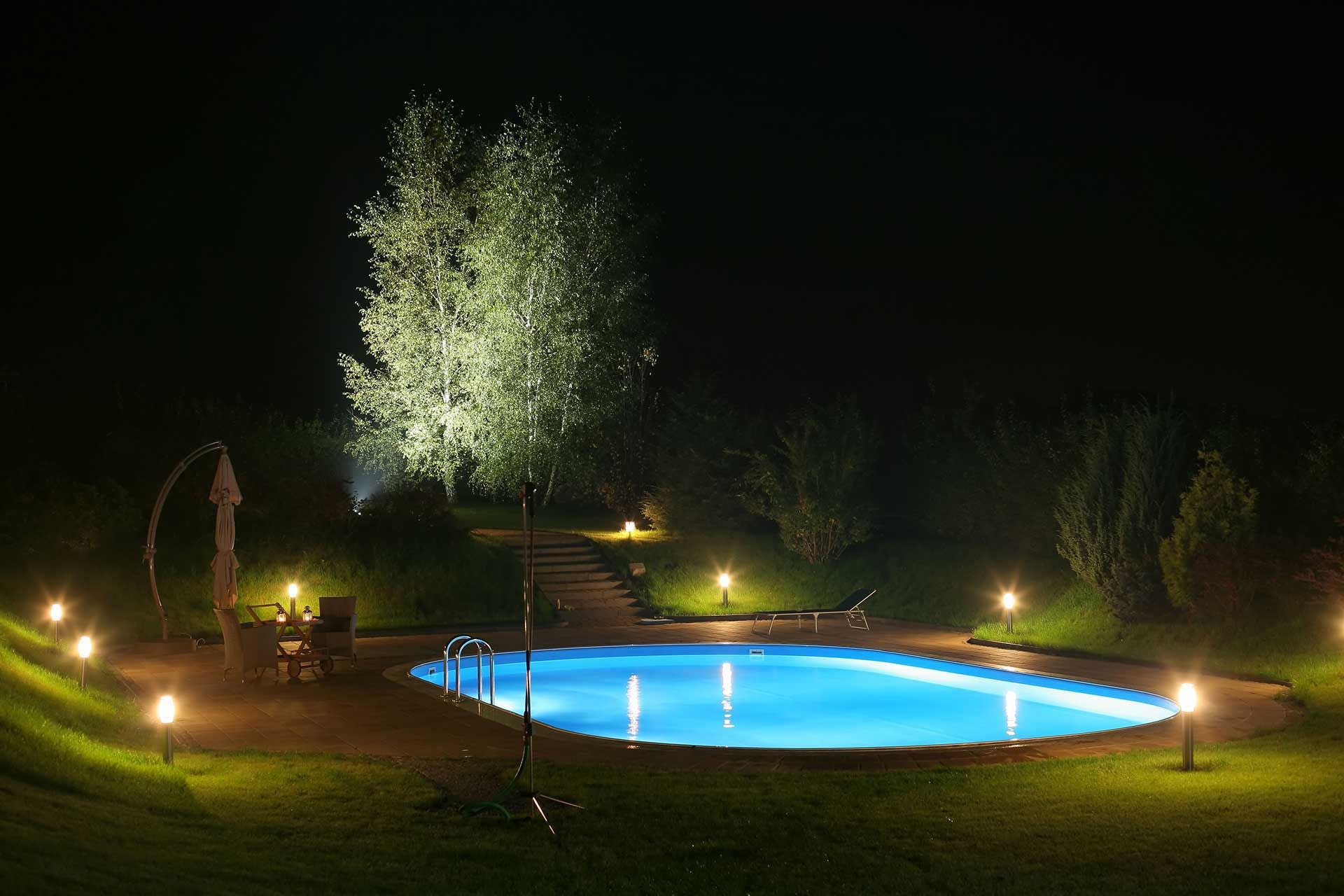 Lit swimming pool at night with lawn lights and a tree in the background