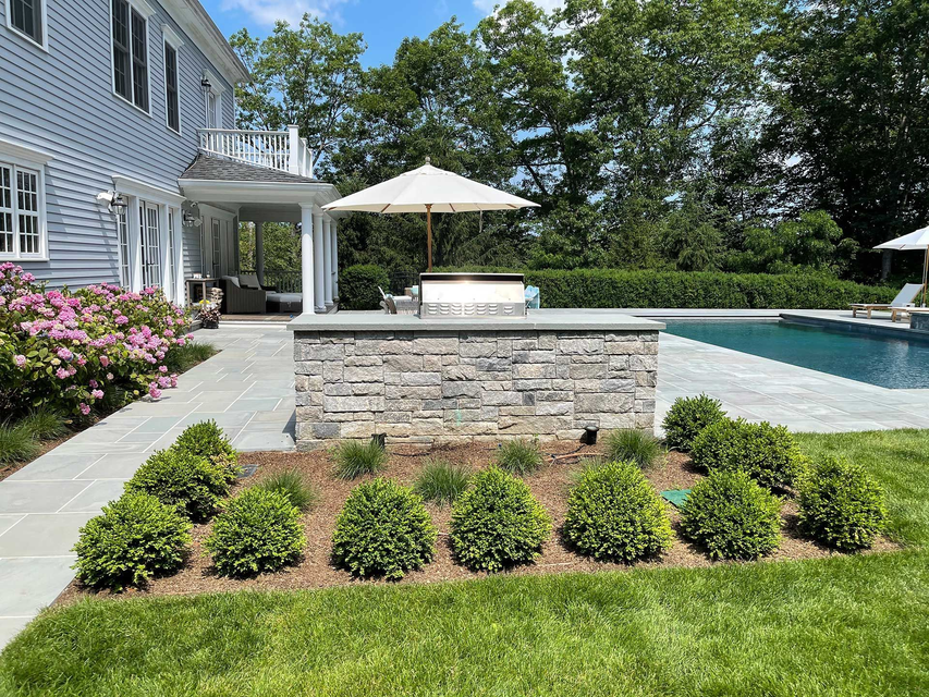 Backyard patio with stone pool wall, umbrella, and house beside a swimming pool and landscaped lawn