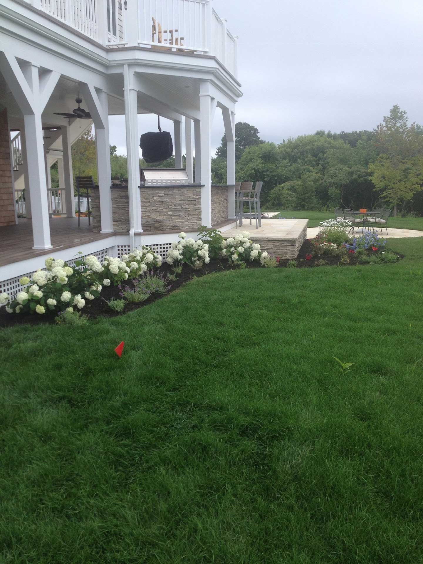 White porch with stone base beside a lush green lawn and flower bed overlooking a misty wooded landscape