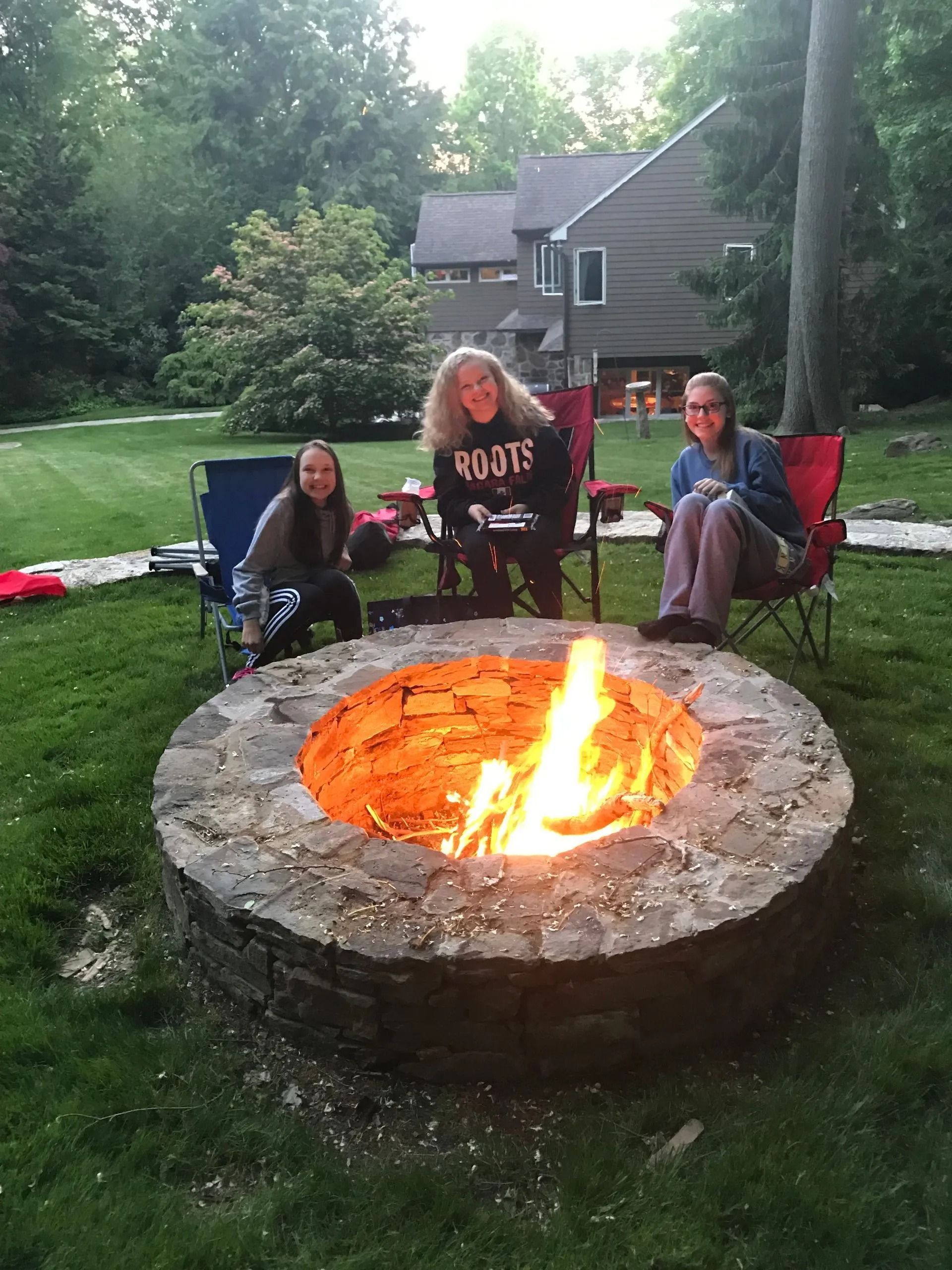 People sitting around a stone fire pit with a bright campfire in a wooded backyard