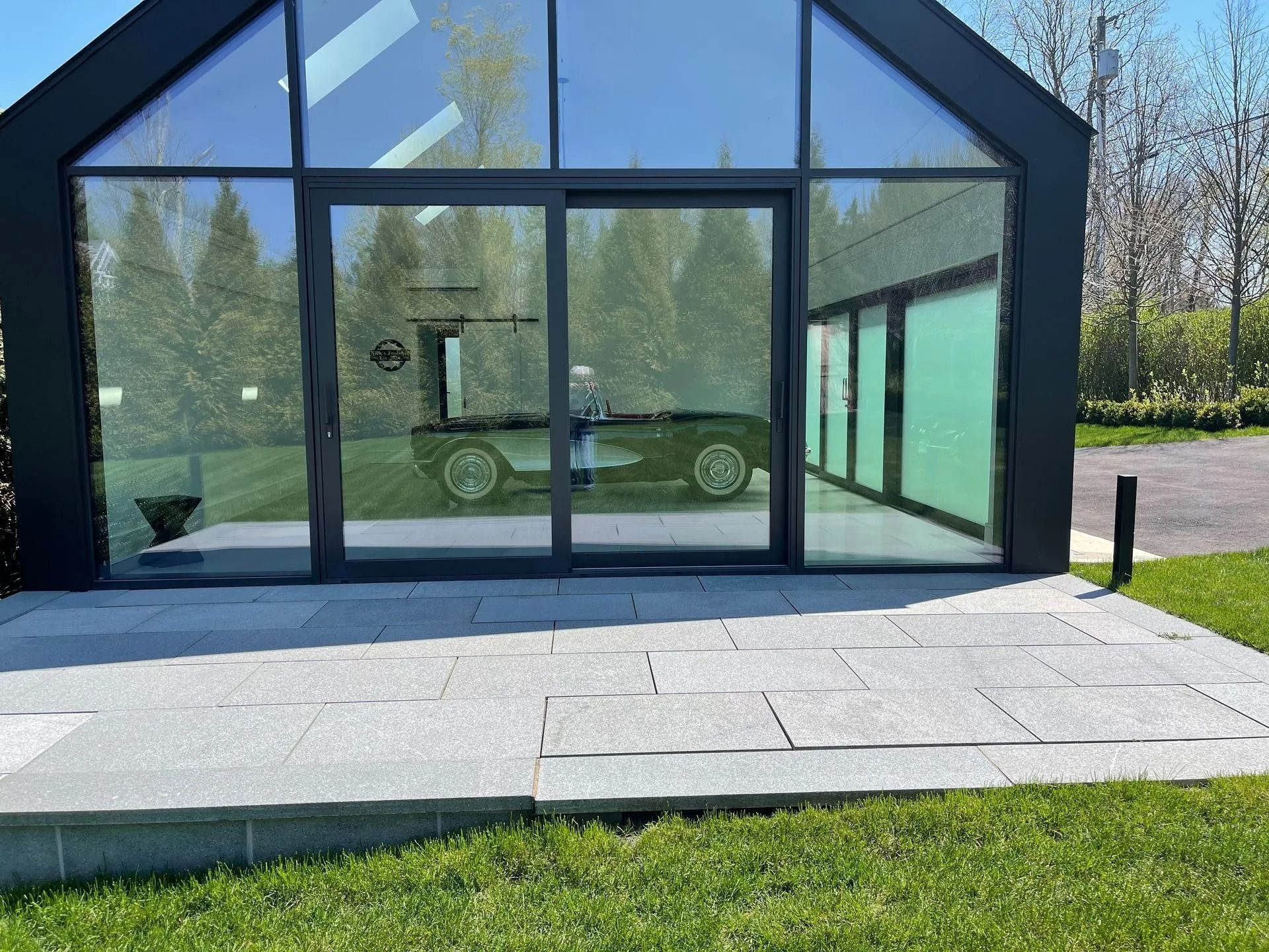 Modern glass-front pavilion with black frame, paved patio, and green lawn in front
