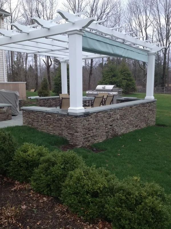 White pergola over stone patio with outdoor seating beside a green lawn and bare trees