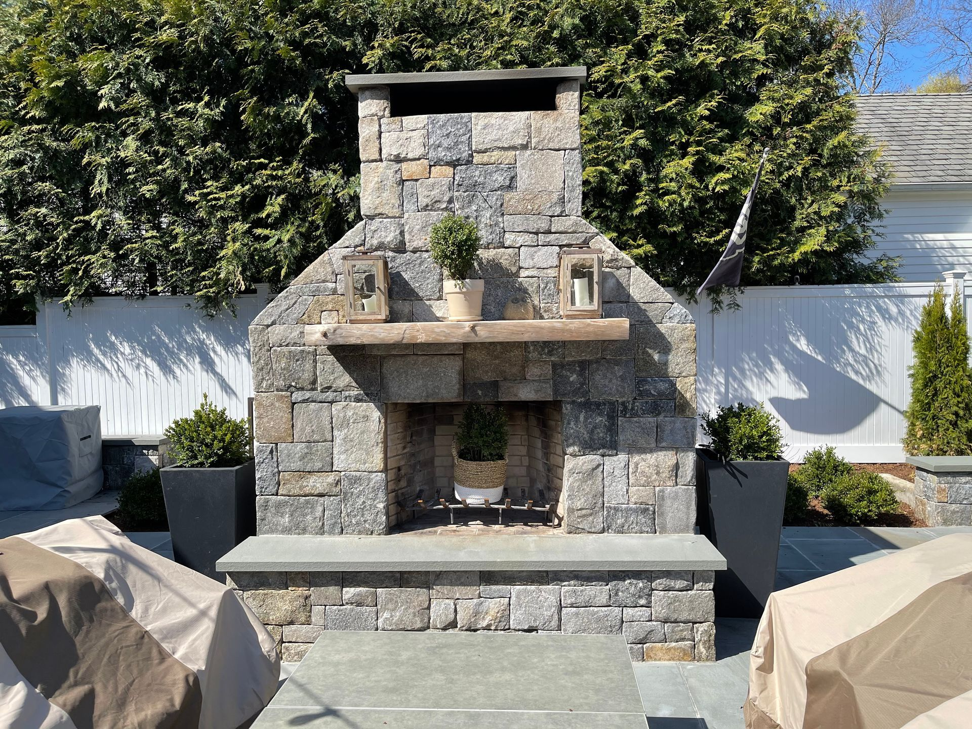 Stone outdoor fireplace with a potted plant, surrounded by covered patio seating and greenery