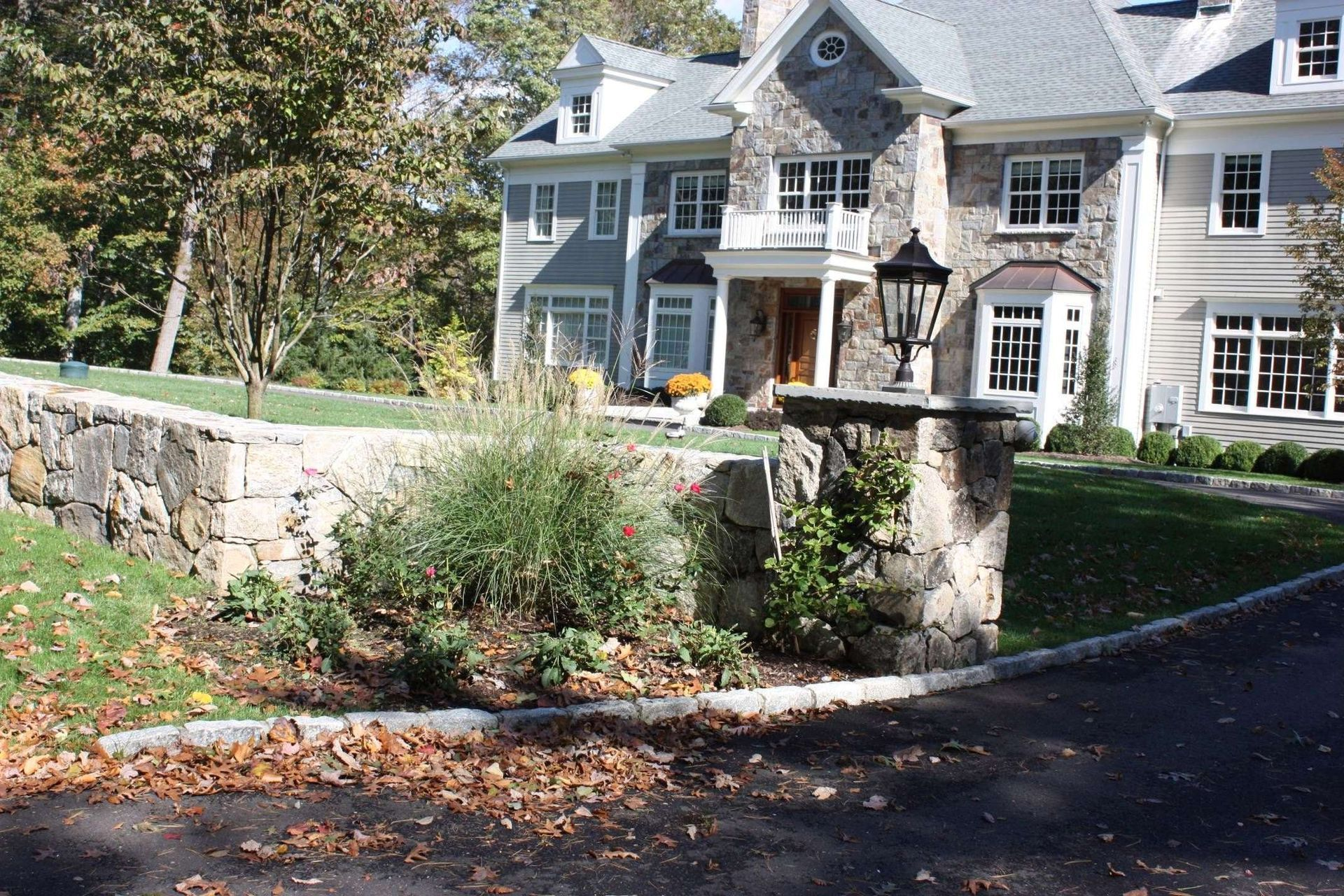Large gray house with white trim and stone wall beside a tree-lined driveway in autumn.