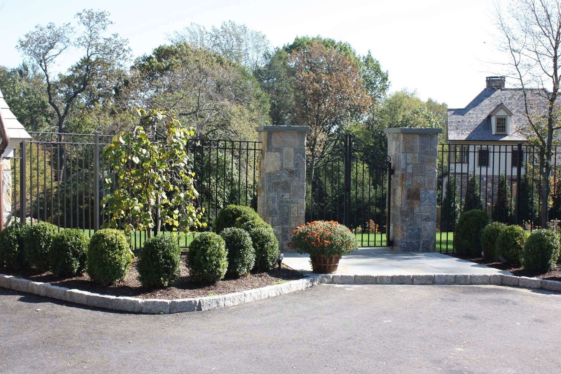 Gated driveway entrance with stone pillars, trimmed shrubs, and trees in front of a house