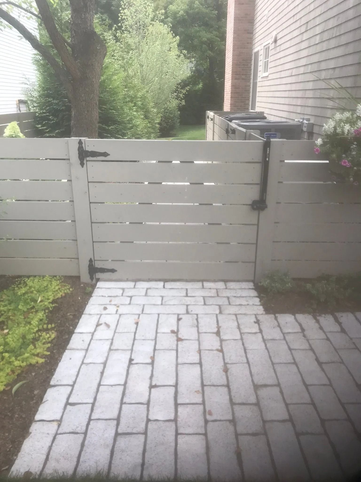 White backyard gate with brick patio, white fence, trees, and house wall beside it