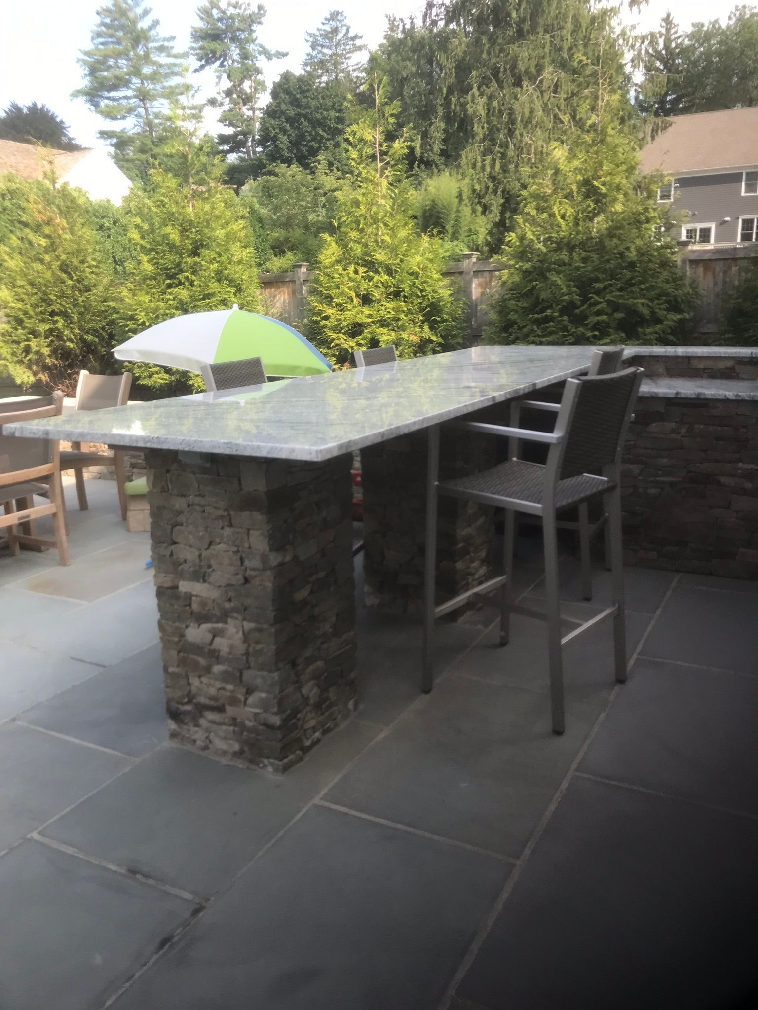 Outdoor patio with stone bar and high chairs, set in a garden with trees and a patio umbrella.