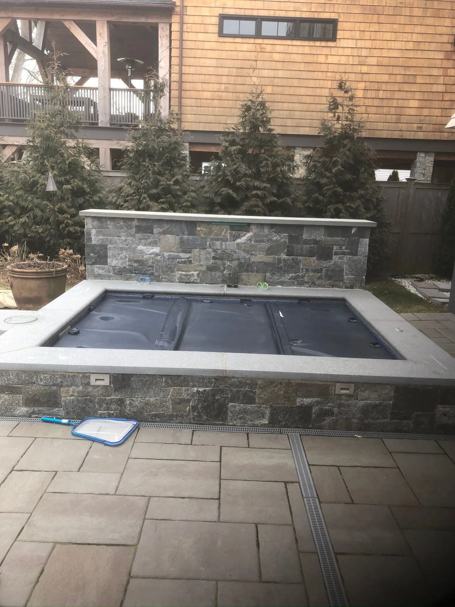 Backyard stone fire pit with black covers, bordered by shrubs and a brick house wall