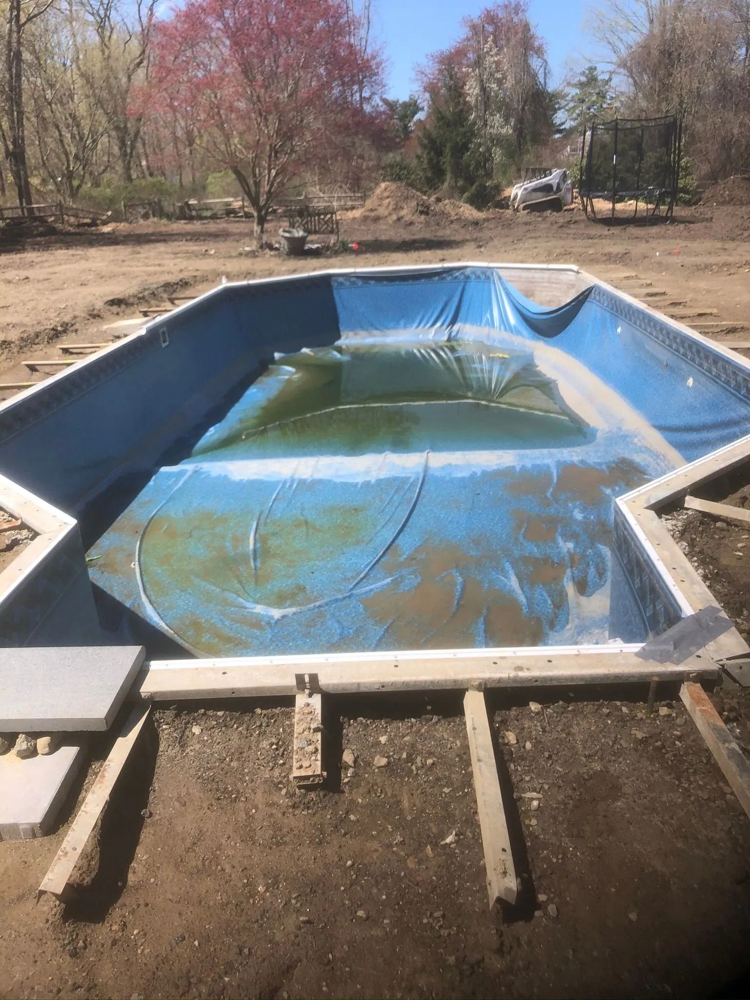 Empty blue in-ground pool with peeling liner in a dry backyard, surrounded by bare trees and dirt.