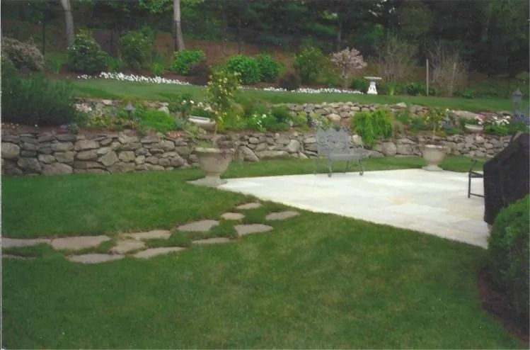 Stone retaining wall with steps beside a green lawn and patio in a landscaped garden.