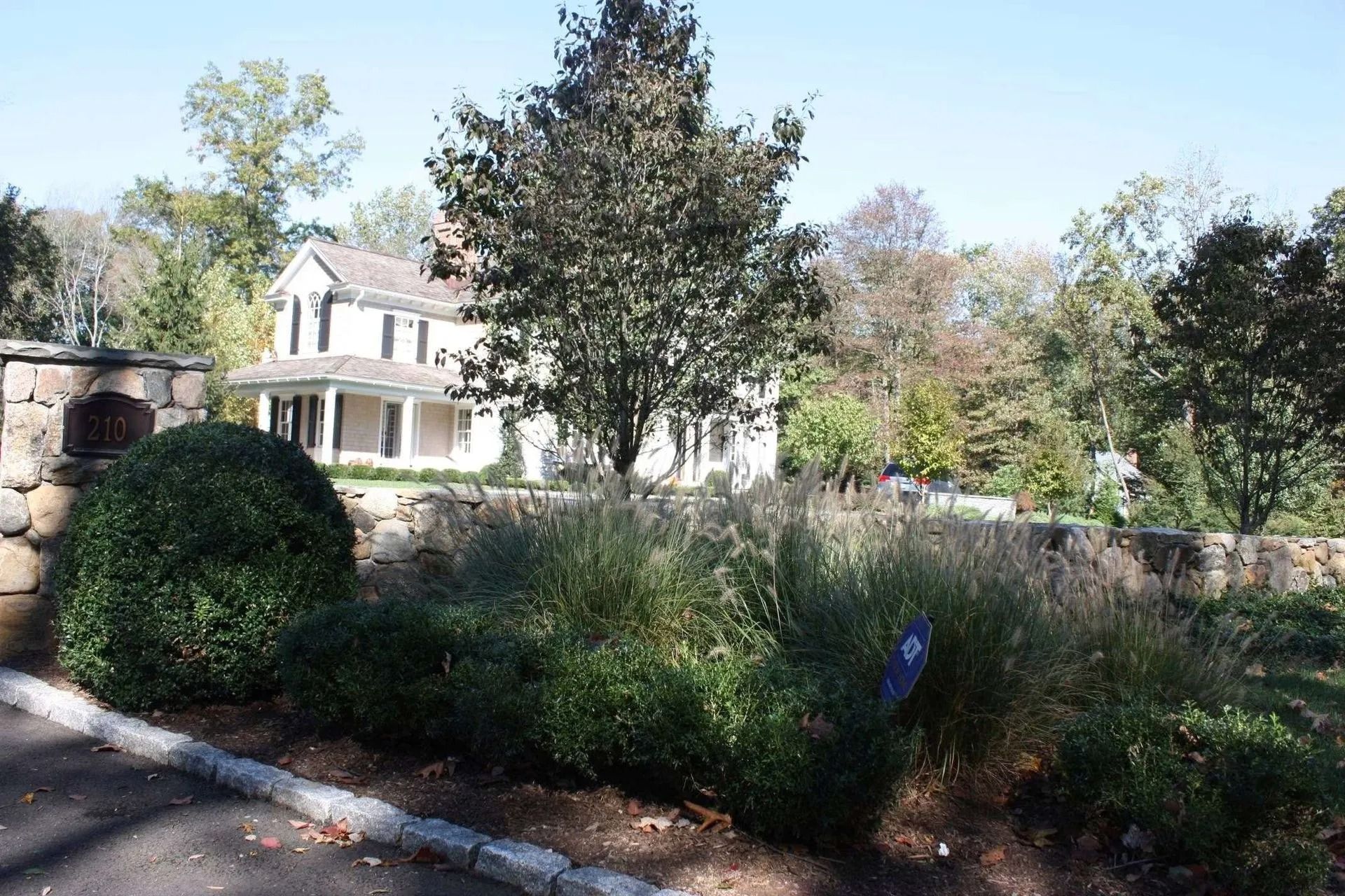 House behind overgrown garden with shrubs, stone wall, and trees in bright daylight